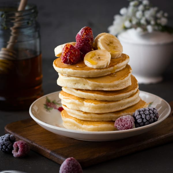 Stack of golden pancakes topped with sliced bananas, raspberries, and honey, with frozen berries nearby.