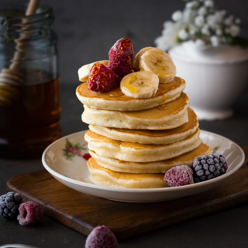 Stack of golden pancakes topped with sliced bananas, raspberries, and honey, with frozen berries nearby.