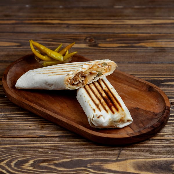 Two grilled wraps, one cut open to show filling, with pickled peppers on a wooden board.