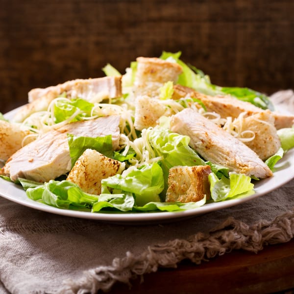Close-up of a Caesar salad with grilled chicken, croutons, and shredded cheese on a white plate.